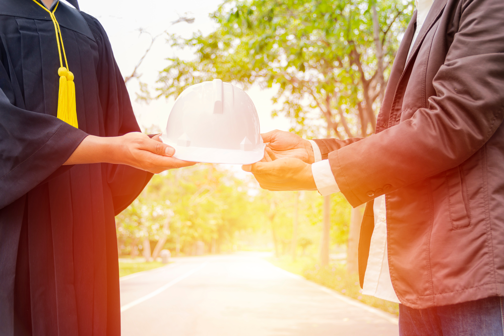 Individual in suit coat passes a hard hat to a person in a graduation gown.