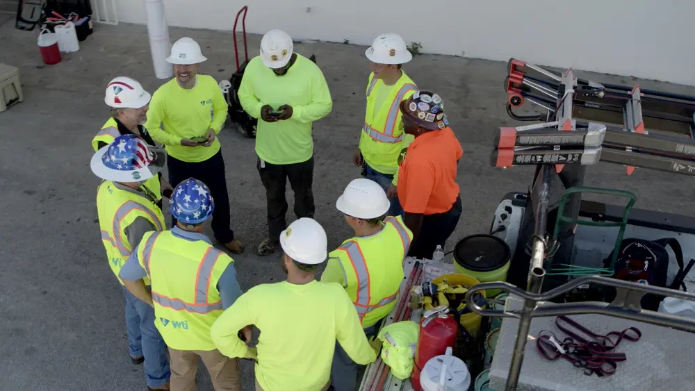 Group of construction workers wearing hard hats and safety gear talk in circle next to truck