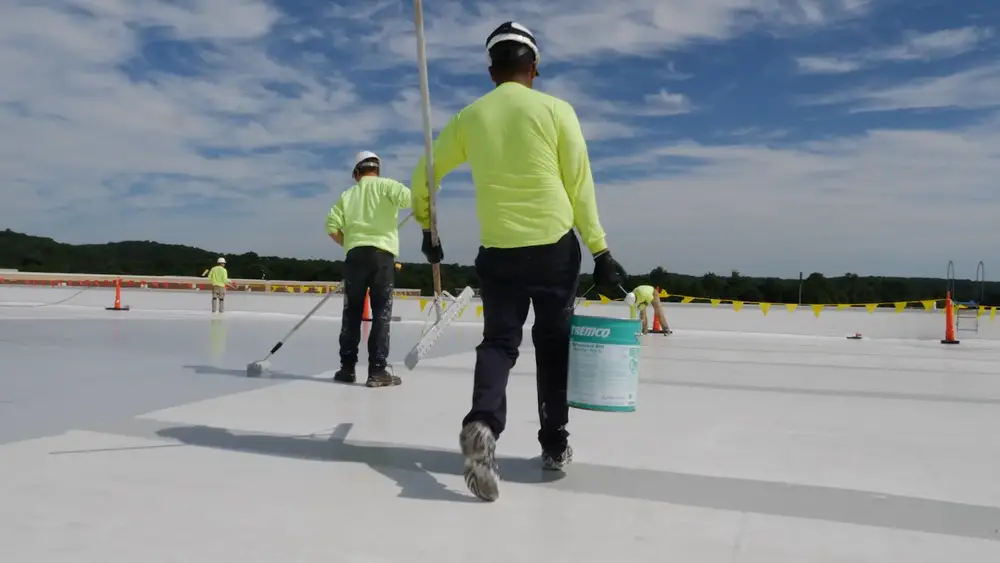 Construction worker carrying bucket on rooftop while workers roll Tremco waterproofing in the background  