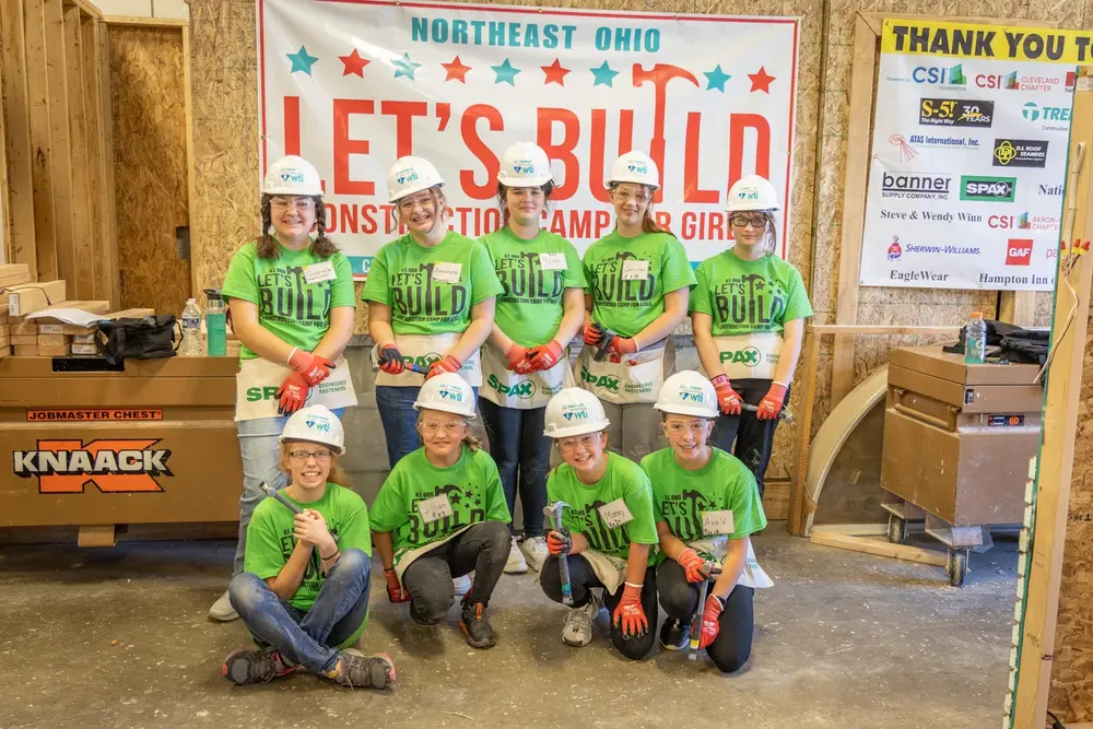 9 girls with hard hats smile in front of at Let’s Build construction camp sign. 