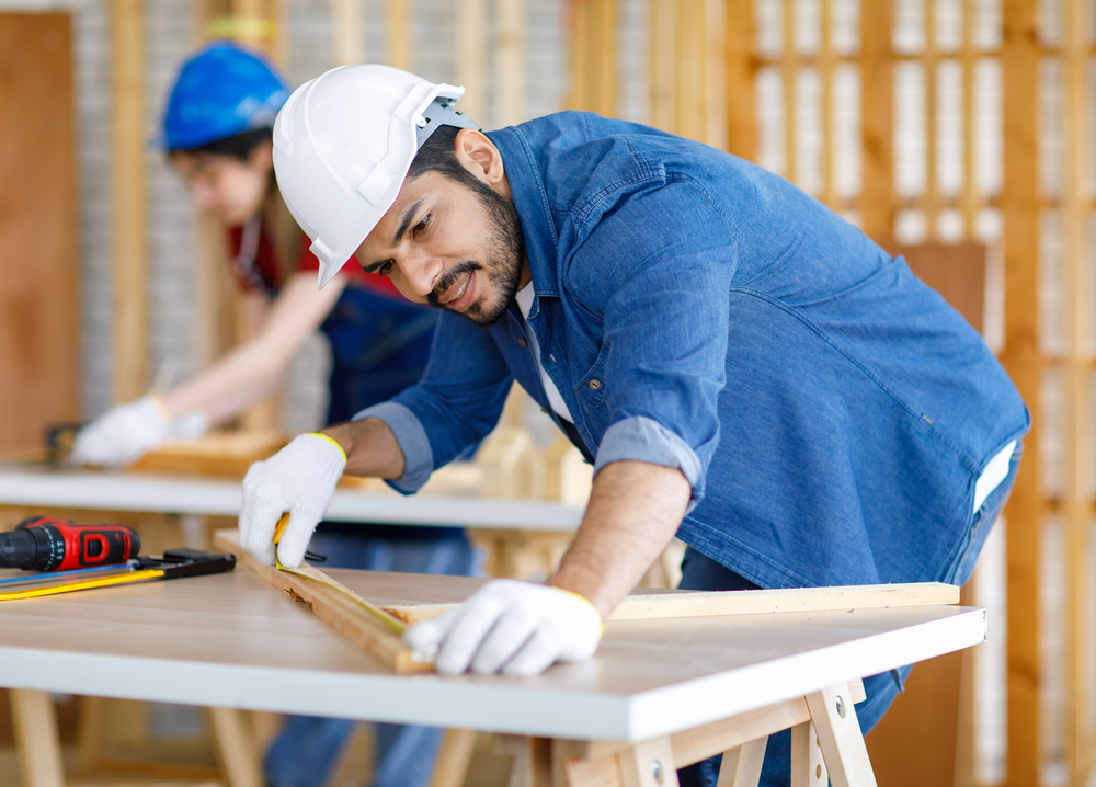 Construction worker in hard hat measuring pieces of wood with tape measure. 